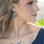 A woman with long blonde hair wearing a blue top and the Holly Yashi Isla Forever Blue Necklace looks to the right in soft natural light.