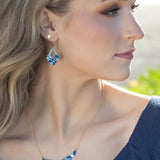 A woman with long blonde hair wearing a blue top and the Holly Yashi Isla Forever Blue Necklace looks to the right in soft natural light.