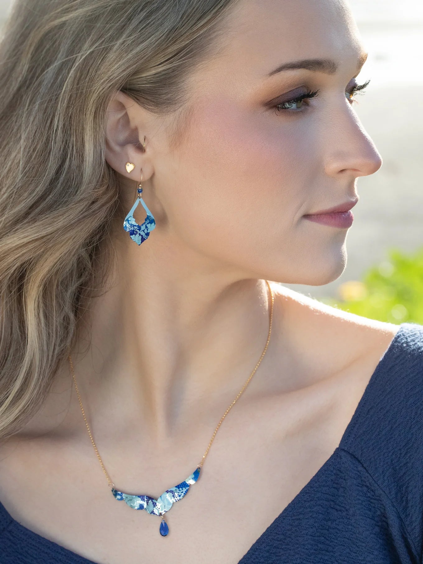 A woman with long blonde hair wearing a blue top and the Holly Yashi Isla Forever Blue Necklace looks to the right in soft natural light.