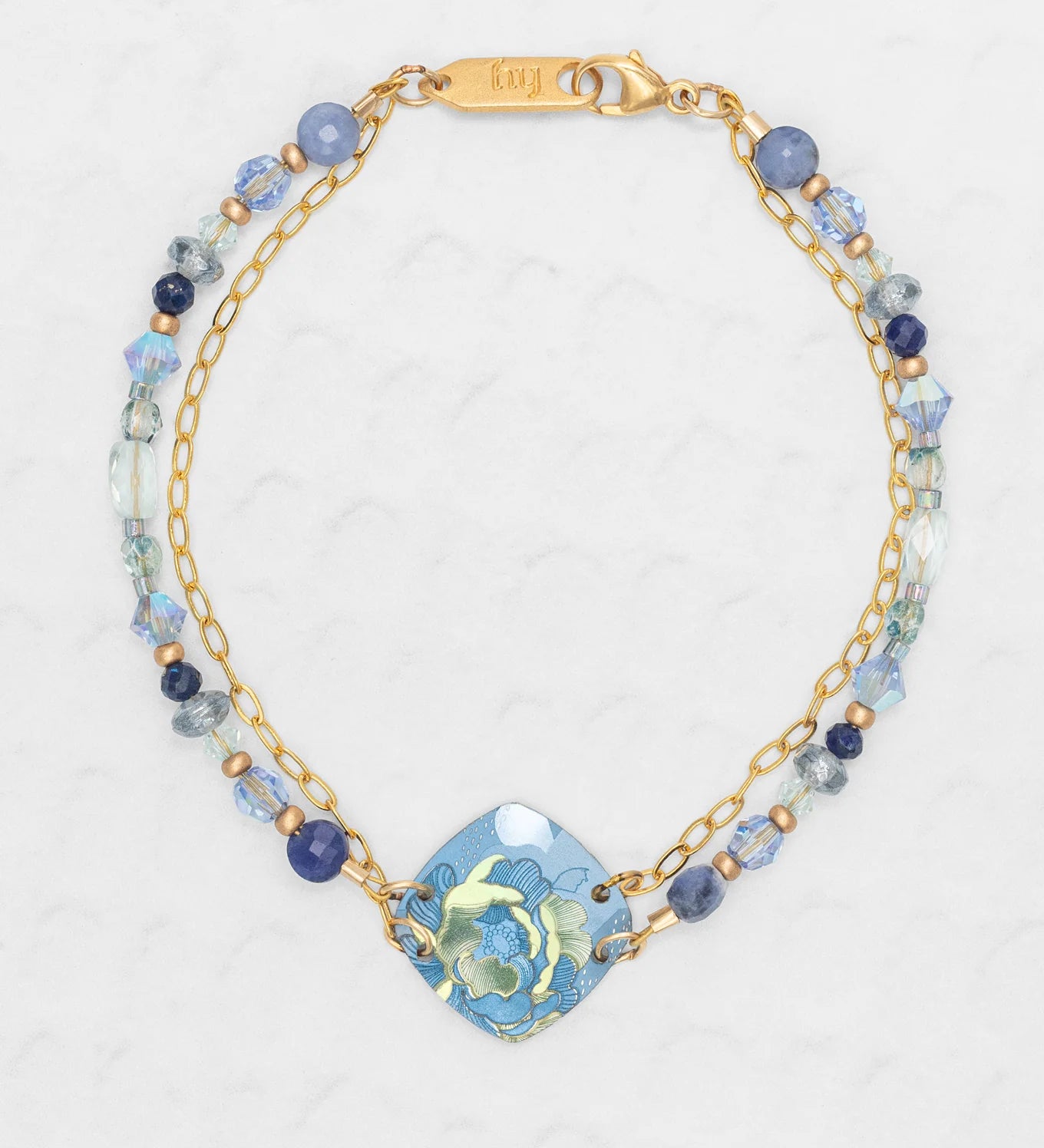 The Elena Beaded Bracelet Sky Blue by Holly Yashi features two gold chains, blue and clear beads, and a blue ceramic floral centerpiece, all displayed on a light textured background.