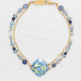 The Elena Beaded Bracelet Sky Blue by Holly Yashi features two gold chains, blue and clear beads, and a blue ceramic floral centerpiece, all displayed on a light textured background.