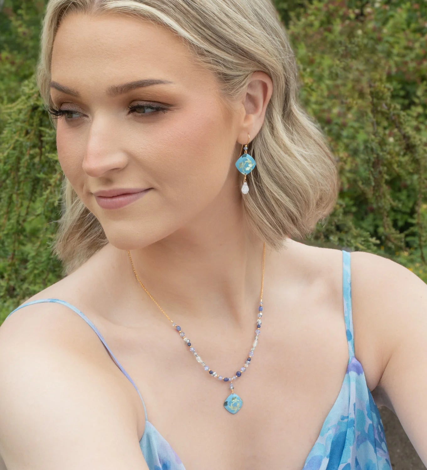A woman with short blonde hair wears the Elena Sky Blue Earrings by Holly Yashi, a blue patterned dress, and a gold necklace with a blue pendant. Outdoors among greenery, she gazes to the side with a gentle smile.