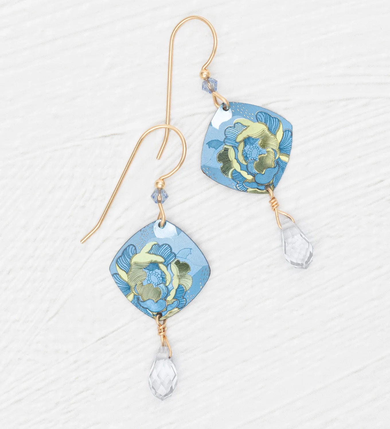 Elena Sky Blue Earrings by Holly Yashi feature gold hooks, blue floral square pendants, and sparkling crystal drops, displayed on a white textured background.