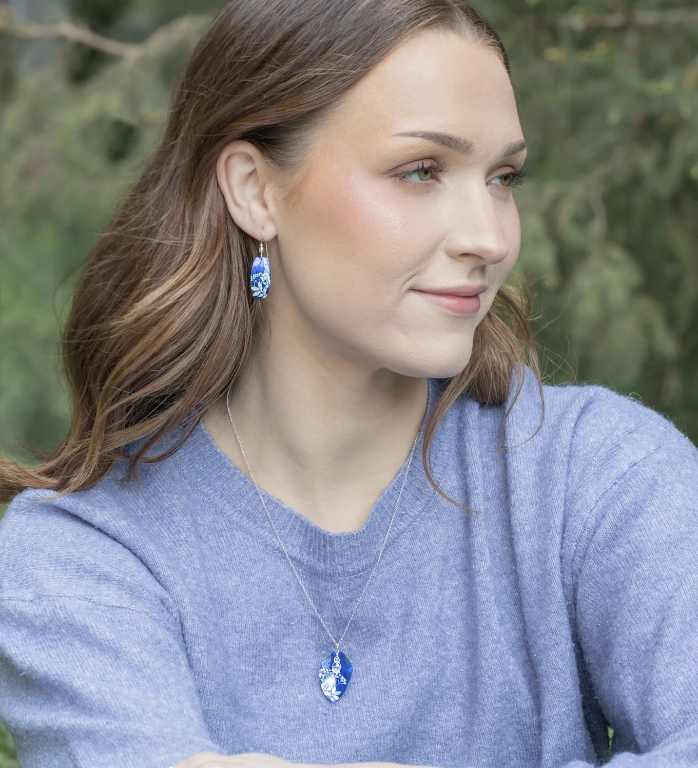 A woman with long brown hair, in a light blue sweater and matching necklace, wears Holly Yashi’s Falling Bloom Indigo Earrings, sitting outdoors against blurred greenery and gazing right with a gentle expression.