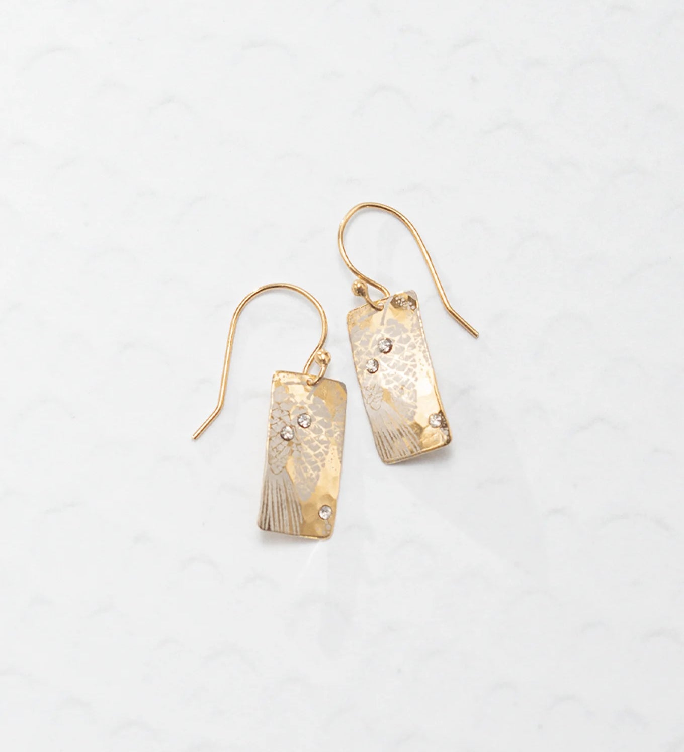 The Radiant Petra Gold Earrings by Holly Yashi are gold-plated with textured rectangular dangles and sparkling gemstone accents, elegantly displayed on a light patterned background.