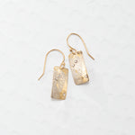 The Radiant Petra Gold Earrings by Holly Yashi are gold-plated with textured rectangular dangles and sparkling gemstone accents, elegantly displayed on a light patterned background.
