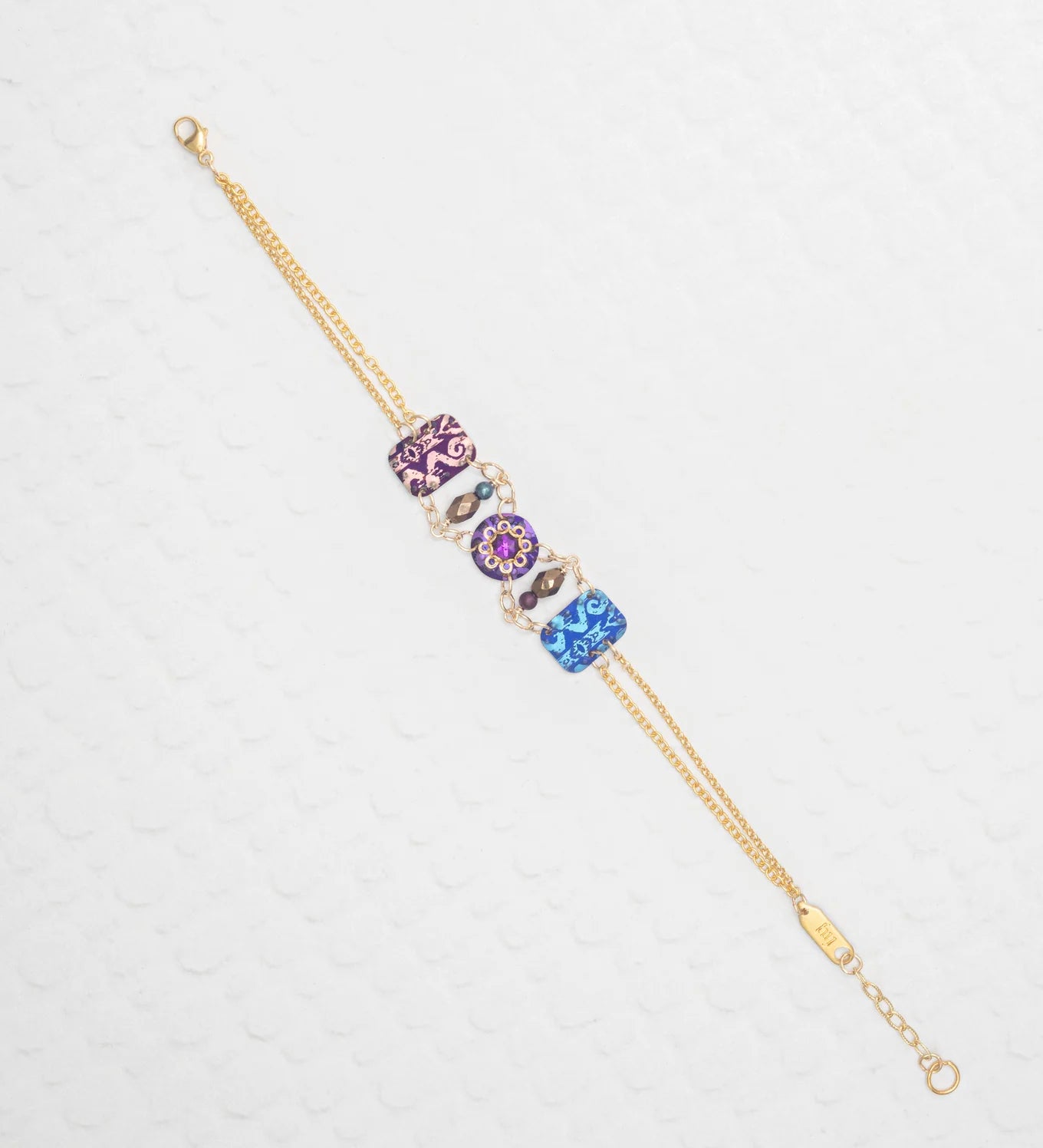 The Alex Multi Color Bracelet by Holly Yashi features a gold chain with three jeweled art charms in purple, pink, and blue, each adorned with floral and swirl patterns, elegantly displayed on a textured white background.