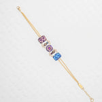 The Alex Multi Color Bracelet by Holly Yashi features a gold chain with three jeweled art charms in purple, pink, and blue, each adorned with floral and swirl patterns, elegantly displayed on a textured white background.