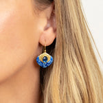 Close-up of a woman wearing Holly Yashi Arabella Sapphire Blue Earrings, which feature a gold and blue design with a central black stone. Her straight, light brown hair partially covers her ear.