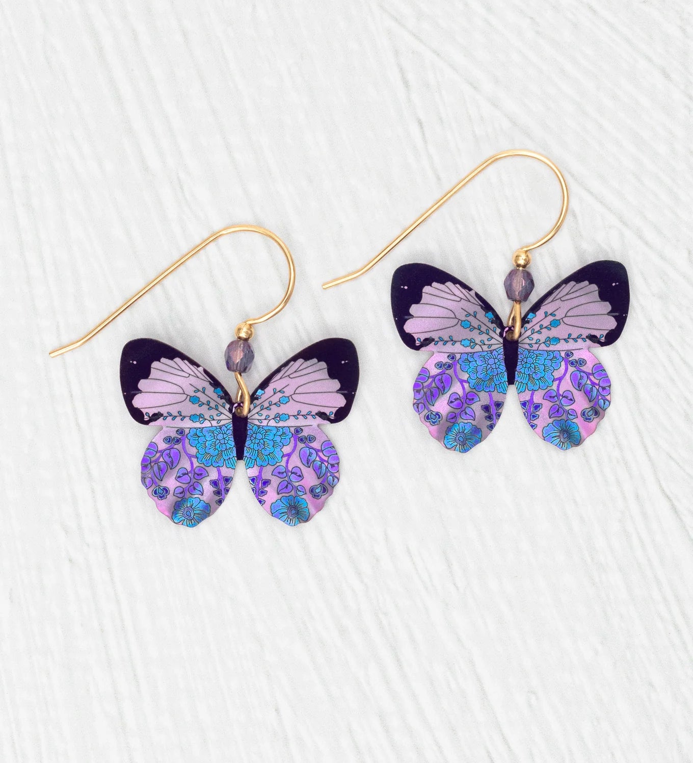Holly Yashi’s ER: Blooming Butterfly Radiant Purple earrings feature gold-filled hooks with intricate butterfly charms in purple, blue, and black patterns, beautifully displayed on a white textured surface.