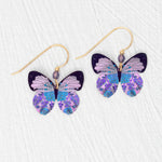 Holly Yashi’s ER: Blooming Butterfly Radiant Purple earrings feature gold-filled hooks with intricate butterfly charms in purple, blue, and black patterns, beautifully displayed on a white textured surface.