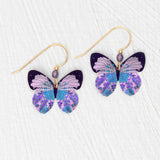 Holly Yashi’s ER: Blooming Butterfly Radiant Purple earrings feature gold-filled hooks with intricate butterfly charms in purple, blue, and black patterns, beautifully displayed on a white textured surface.
