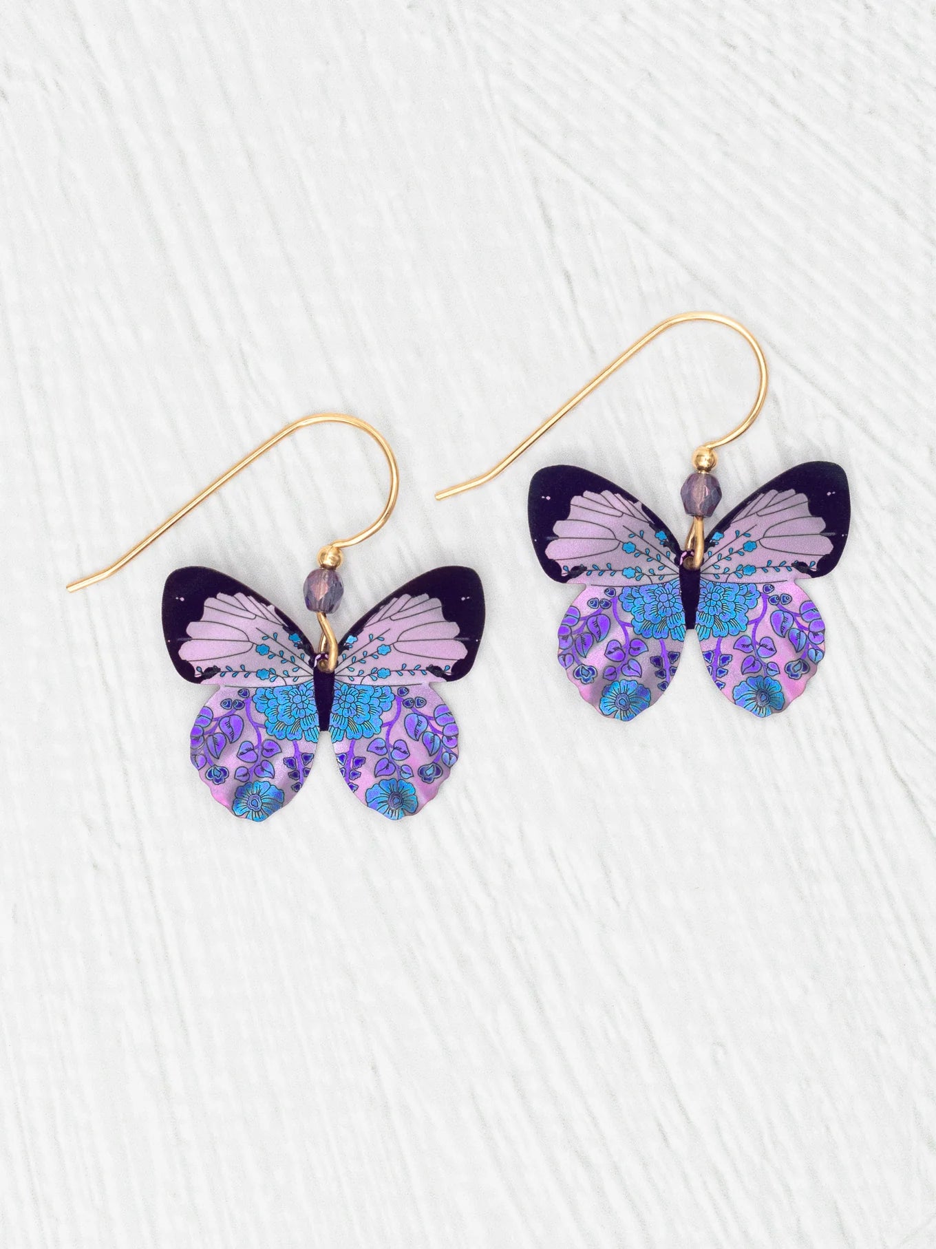 Holly Yashi’s ER: Blooming Butterfly Radiant Purple earrings feature gold-filled hooks with intricate butterfly charms in purple, blue, and black patterns, beautifully displayed on a white textured surface.