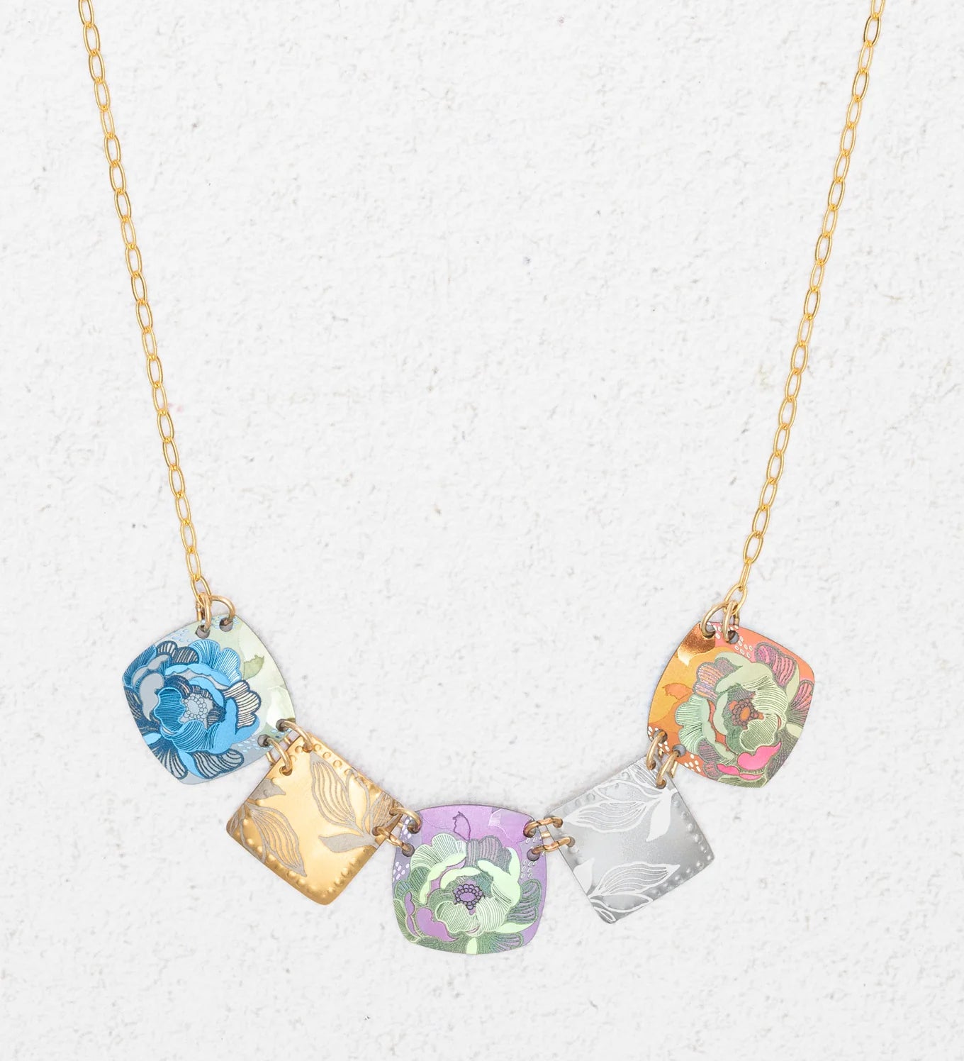 The Flora Multi Color Necklace by Holly Yashi is a handcrafted gold chain with five square pendants in blue, gold, purple, silver, and orange floral patterns, shown on a light textured background.
