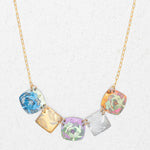 The Flora Multi Color Necklace by Holly Yashi is a handcrafted gold chain with five square pendants in blue, gold, purple, silver, and orange floral patterns, shown on a light textured background.