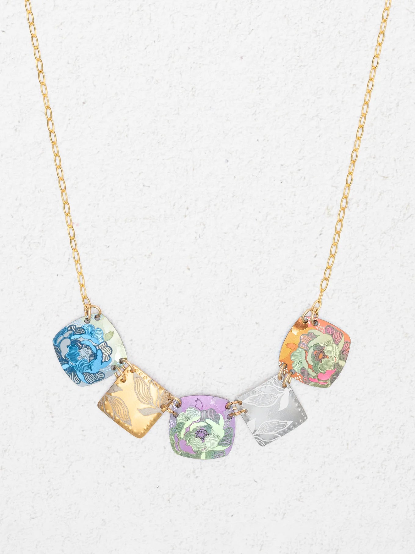The Flora Multi Color Necklace by Holly Yashi is a handcrafted gold chain with five square pendants in blue, gold, purple, silver, and orange floral patterns, shown on a light textured background.