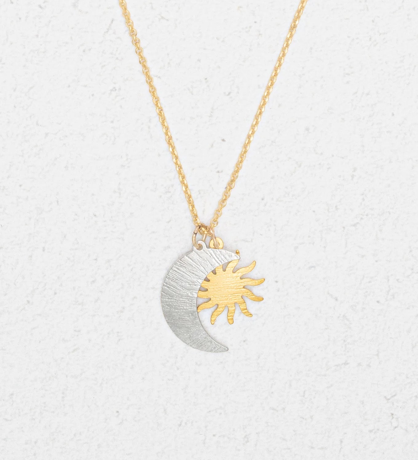 The Holly Yashi Sun and Moon Gold & Silver Necklace features overlapping silver crescent moon and gold sun pendants, reflecting a celestial jewelry style, displayed on a white textured background.