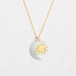The Holly Yashi Sun and Moon Gold & Silver Necklace features overlapping silver crescent moon and gold sun pendants, reflecting a celestial jewelry style, displayed on a white textured background.