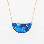The Holly Yashi Orion Pendant/ Midnight Blue Necklace features a half-circle pendant with subtle floral designs and gold accents on a gold chain, displayed against a white textured background.