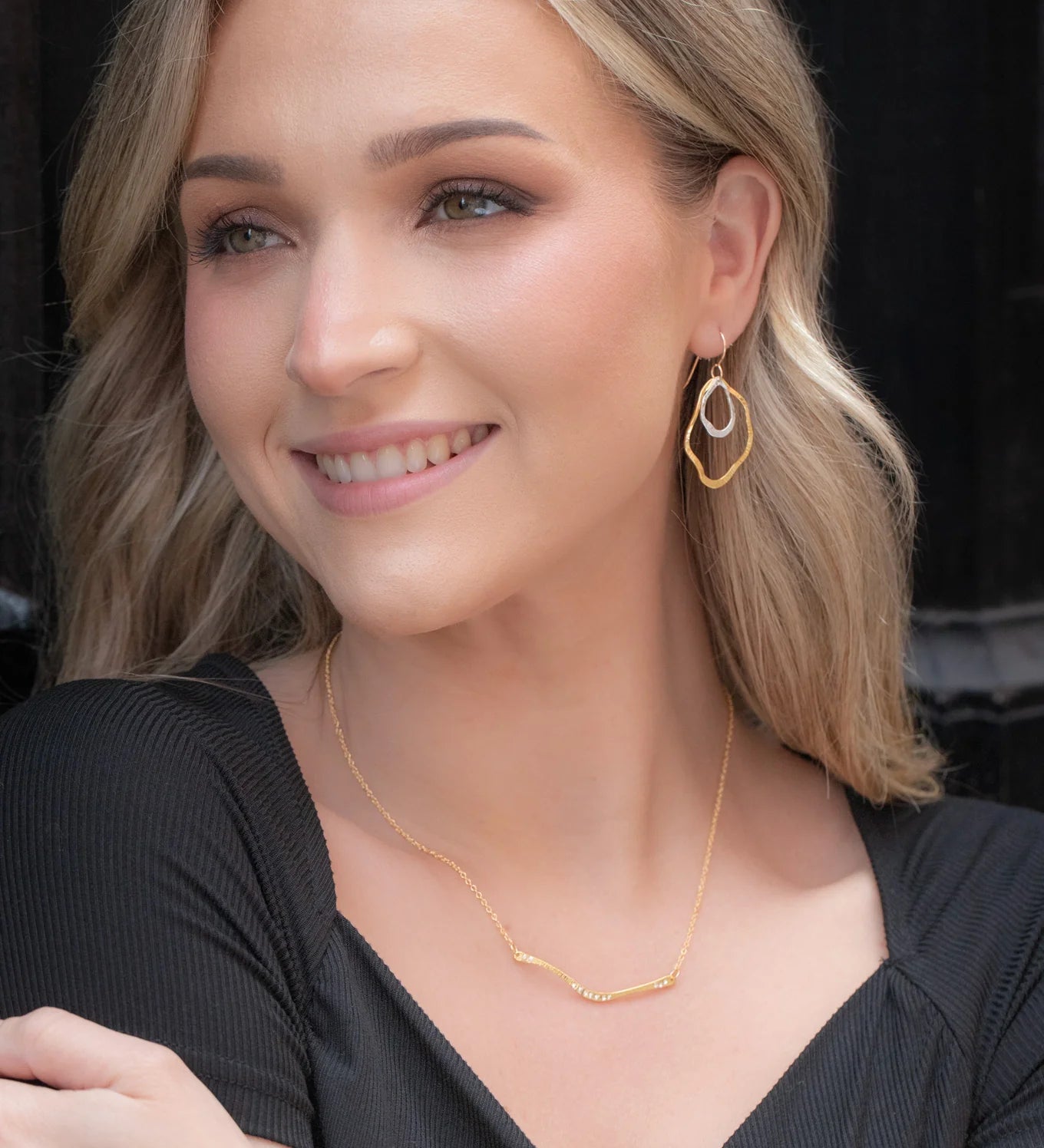 A woman with long blonde hair smiles in a black dress and hypoallergenic gold geometric earrings, wearing the Zara Bar Gold Necklace by Holly Yashi. She stands against a dark background.