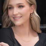 A woman with long blonde hair smiles in a black dress and hypoallergenic gold geometric earrings, wearing the Zara Bar Gold Necklace by Holly Yashi. She stands against a dark background.
