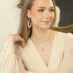 A woman with long, light brown hair wears a cream blouse, layered gold necklaces, and Rafaela Gold & White Earrings by Holly Yashi as she sits with her arm on a marble surface, smiling slightly with a decorative mirror behind her.