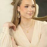 A woman with long, light brown hair wears a cream blouse, layered gold necklaces, and Rafaela Gold & White Earrings by Holly Yashi as she sits with her arm on a marble surface, smiling slightly with a decorative mirror behind her.