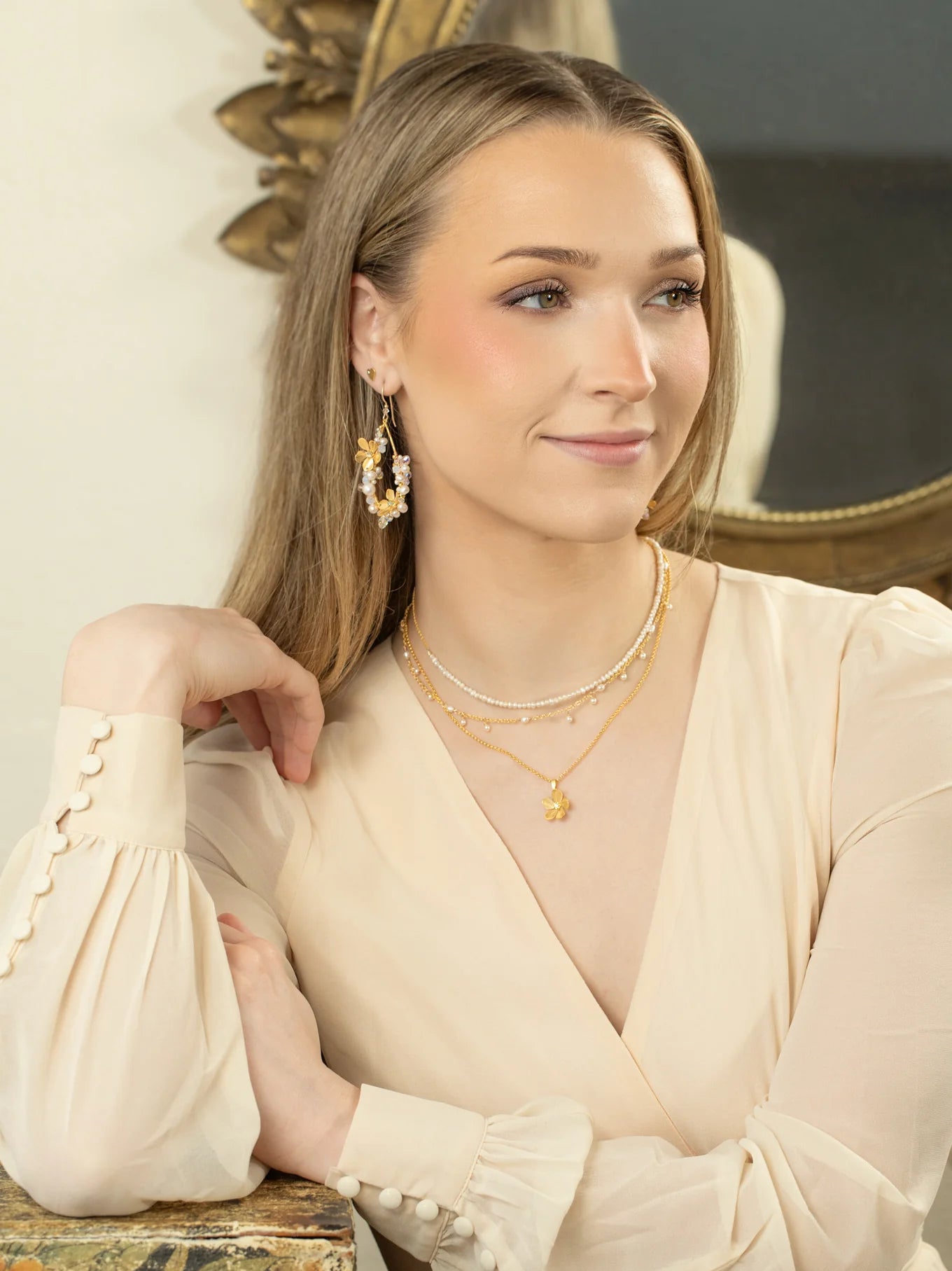 A woman with long, light brown hair wears a cream blouse, layered gold necklaces, and Rafaela Gold & White Earrings by Holly Yashi as she sits with her arm on a marble surface, smiling slightly with a decorative mirror behind her.