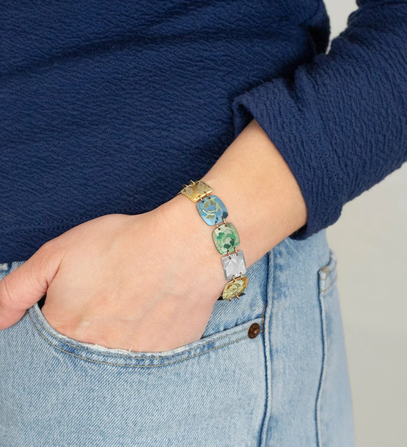 A person wearing a blue long-sleeve top and light blue jeans has their hand in their pocket, showcasing vibrant Flora Multi Color Earrings by Holly Yashi.