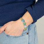 A person wearing a blue long-sleeve top and light blue jeans has their hand in their pocket, showcasing vibrant Flora Multi Color Earrings by Holly Yashi.