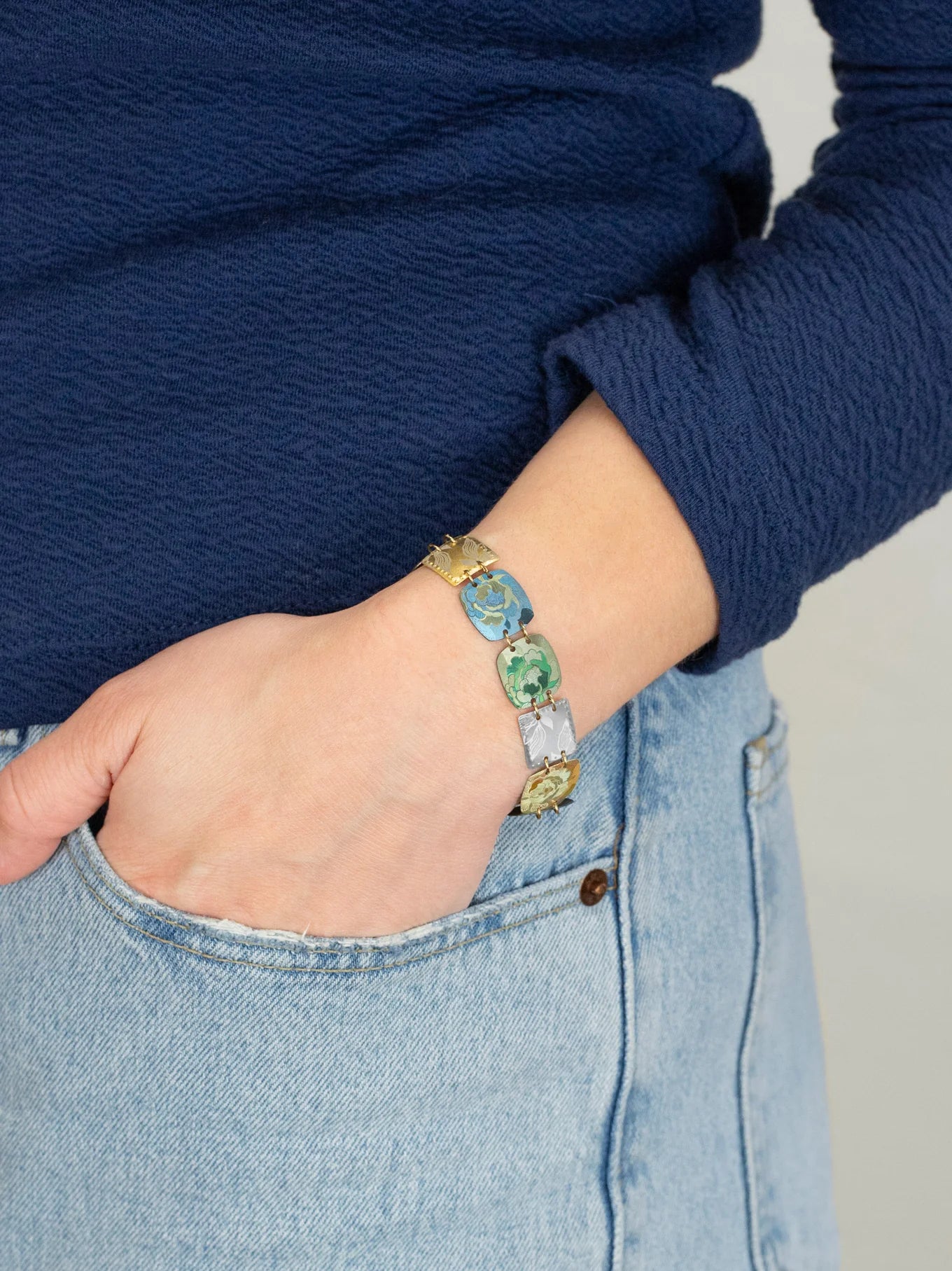 A person wearing a blue long-sleeve top and light blue jeans has their hand in their pocket, showcasing vibrant Flora Multi Color Earrings by Holly Yashi.