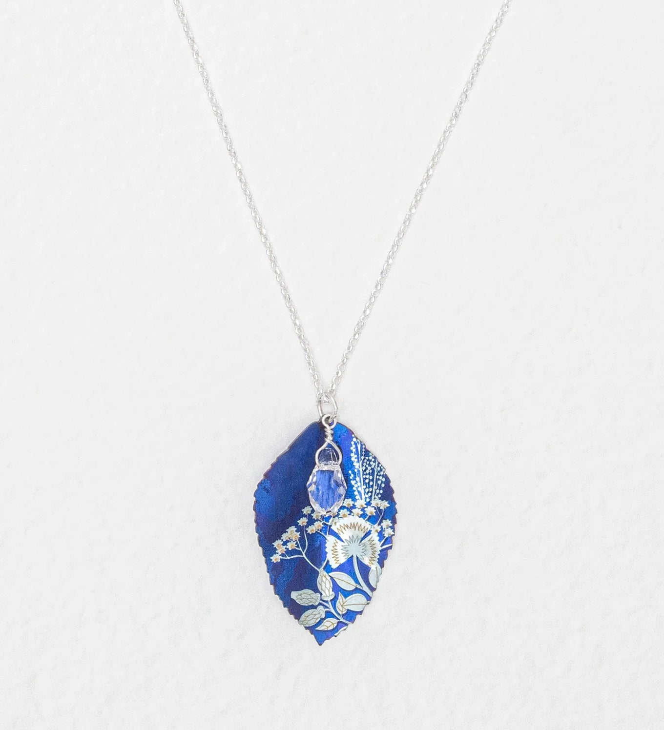 The Blooming Elm Indigo Necklace by Holly Yashi features a silver chain and a blue, leaf-shaped pendant with intricate white botanical designs. This elegant piece of jewelry is shown against a white background.