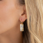 Close-up of a woman's ear wearing the Radiant Petra Gold Earrings by Holly Yashi—gold-plated dangle earrings with a textured metallic finish. Her blonde hair partly frames her ear, accentuating these elegant statement pieces.