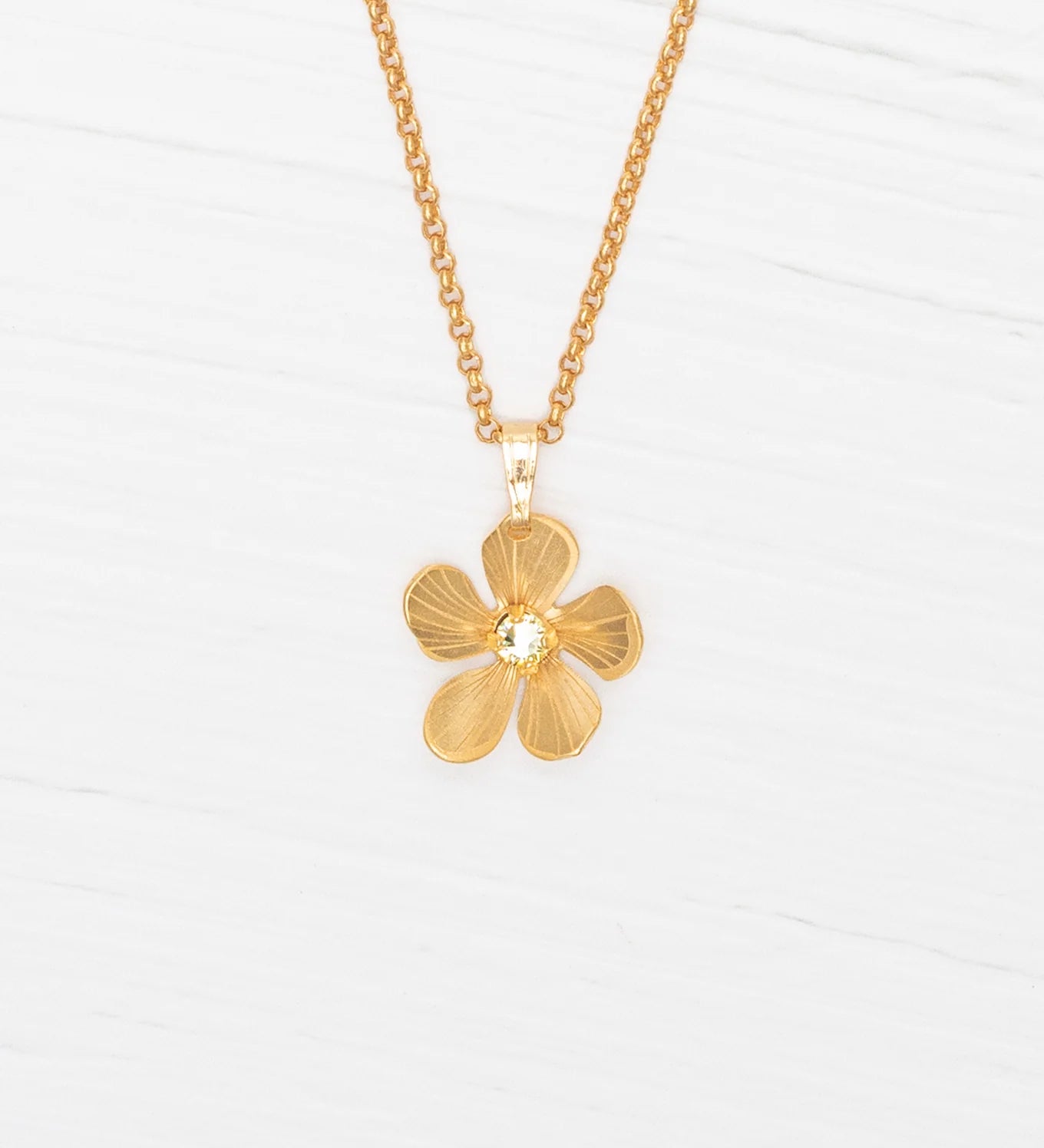 The Plumeria Drop Gold Necklace by Holly Yashi features a hand-formed five-petal bloom pendant on a delicate chain, beautifully displayed on a white wooden surface.