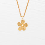 The Plumeria Drop Gold Necklace by Holly Yashi features a hand-formed five-petal bloom pendant on a delicate chain, beautifully displayed on a white wooden surface.