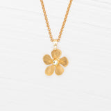 The Plumeria Drop Gold Necklace by Holly Yashi features a hand-formed five-petal bloom pendant on a delicate chain, beautifully displayed on a white wooden surface.