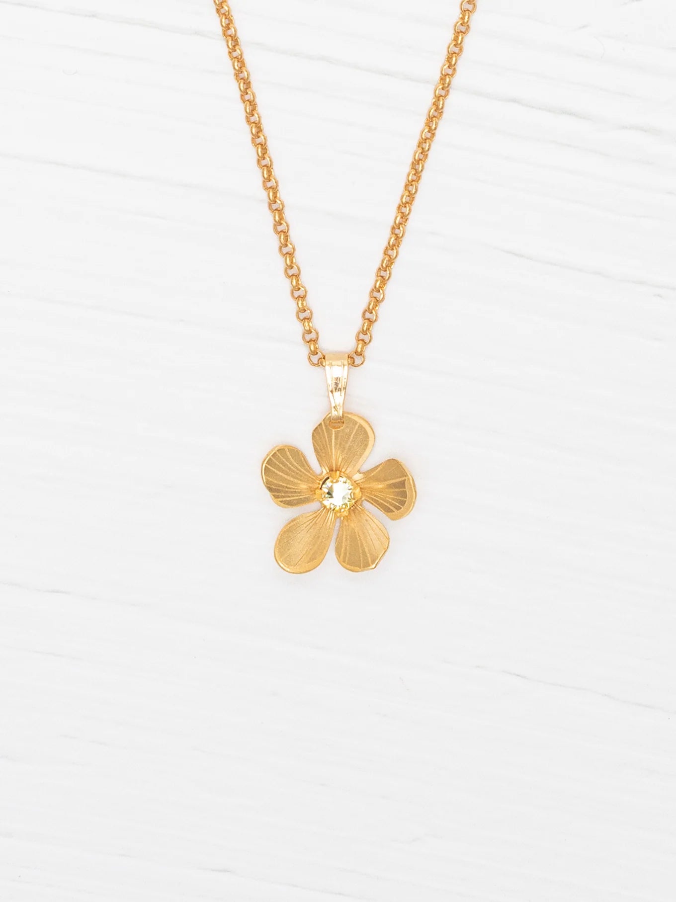 The Plumeria Drop Gold Necklace by Holly Yashi features a hand-formed five-petal bloom pendant on a delicate chain, beautifully displayed on a white wooden surface.