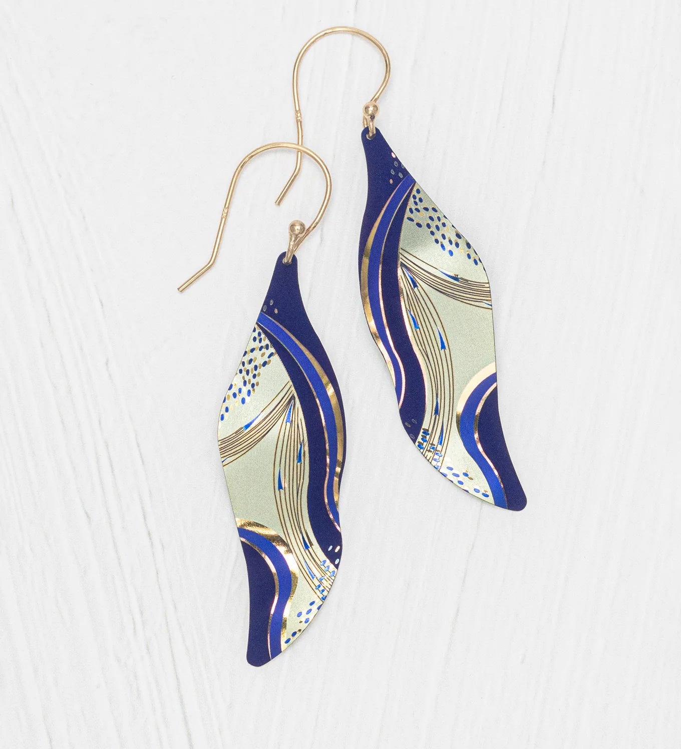 The Turning Leaf Indigo Earrings by Holly Yashi feature wavy, leaf-shaped drops with a blue, gold, and cream abstract design on gold-filled wires, shown on a white textured background.