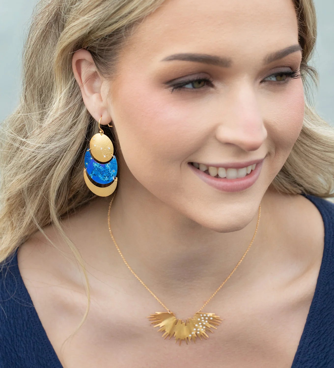 A woman with wavy blonde hair smiles, wearing a navy top and the Holly Yashi Aster Necklace-Gold Necklace, which features a sunburst design, along with large gold dangle earrings accented with shimmering blue crystal stones.