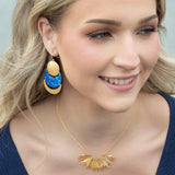 A woman with wavy blonde hair smiles, wearing a navy top and the Holly Yashi Aster Necklace-Gold Necklace, which features a sunburst design, along with large gold dangle earrings accented with shimmering blue crystal stones.