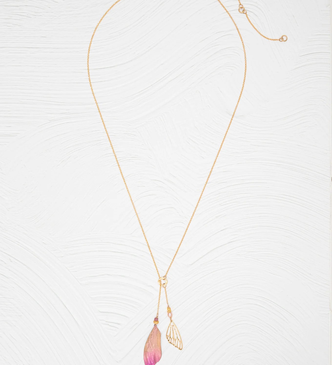 The Holly Yashi Special Edition Flutterby Pink Lariat Necklace features a fine gold chain with two pendants—a metallic leaf and a pink-tipped, translucent leaf—displayed on a white textured background.