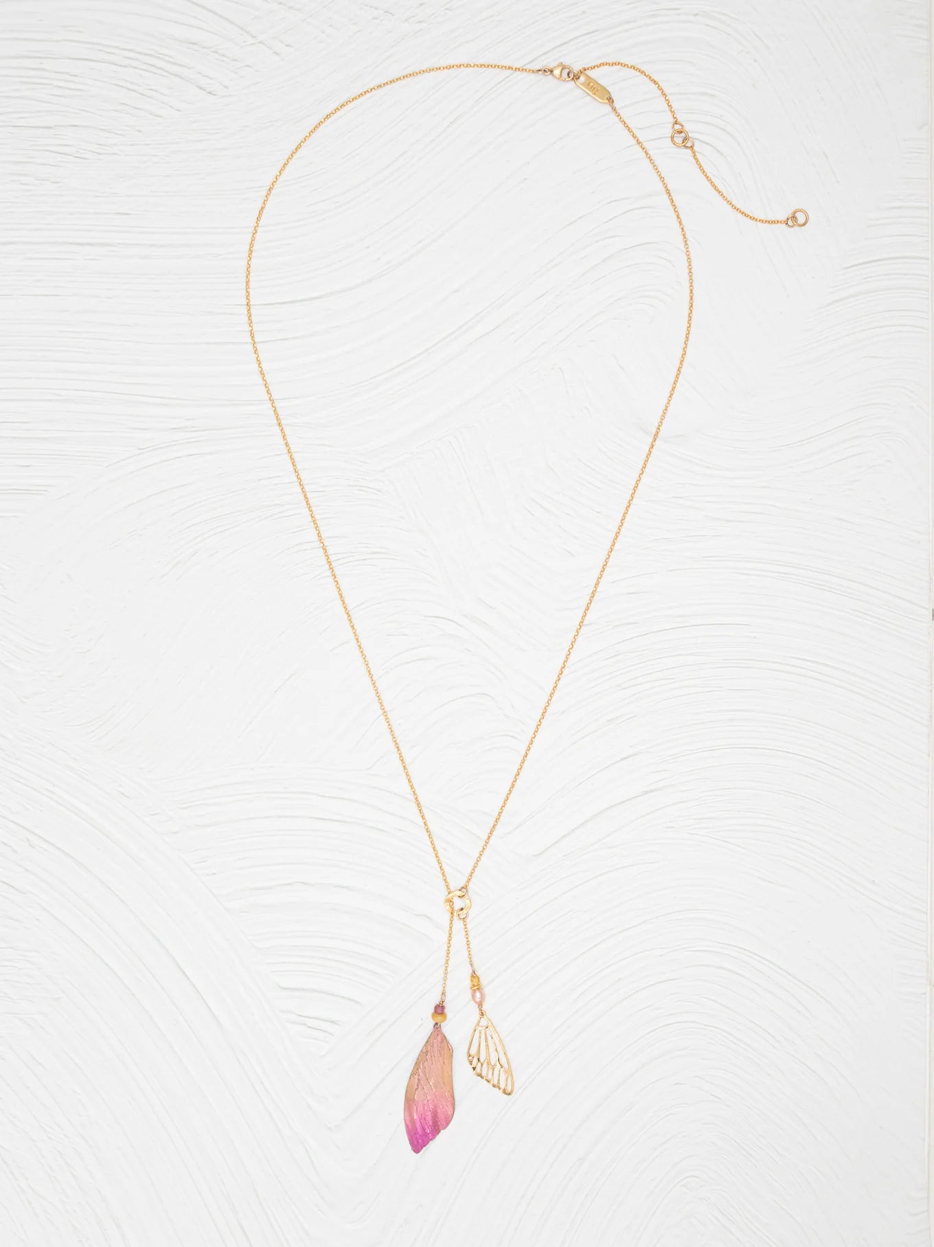 The Holly Yashi Special Edition Flutterby Pink Lariat Necklace features a fine gold chain with two pendants—a metallic leaf and a pink-tipped, translucent leaf—displayed on a white textured background.