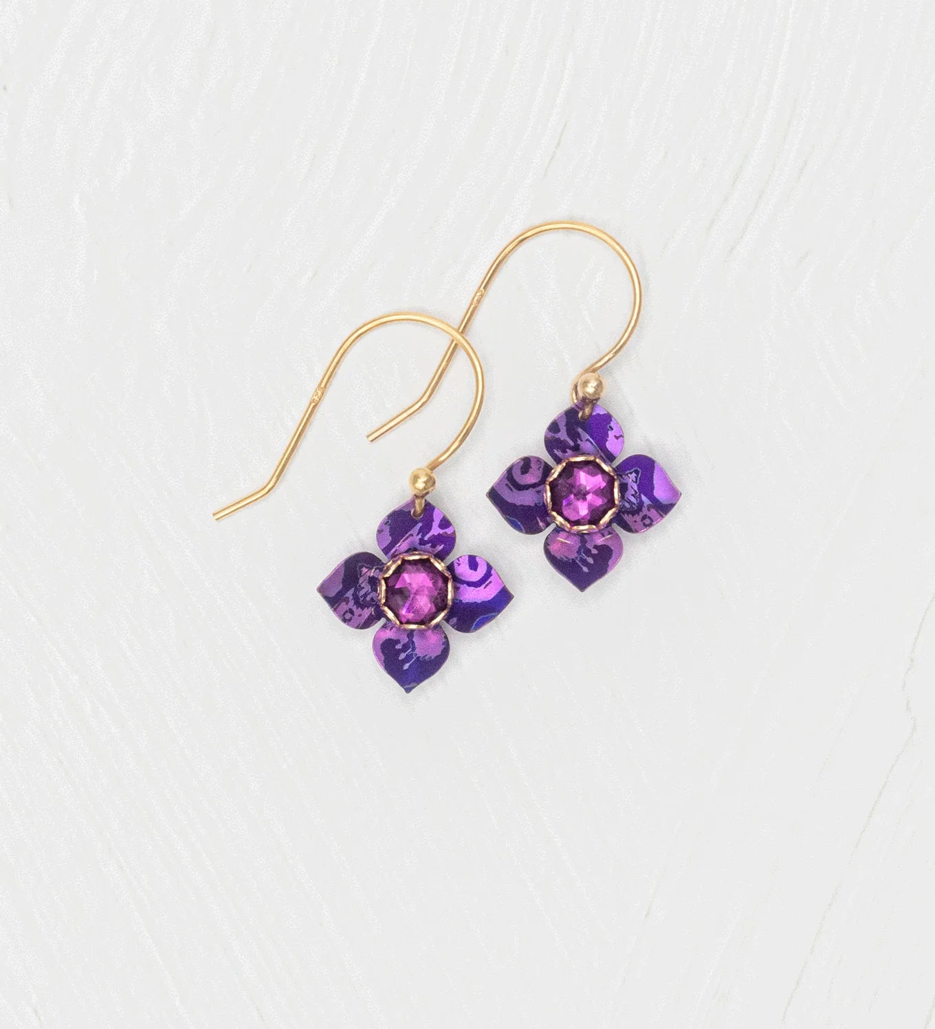 The Holly Yashi Petite Fleur Royal Purple Earrings feature gold hooks with purple flower-shaped pendants and faceted Austrian crystal centers, shown on a white textured background.