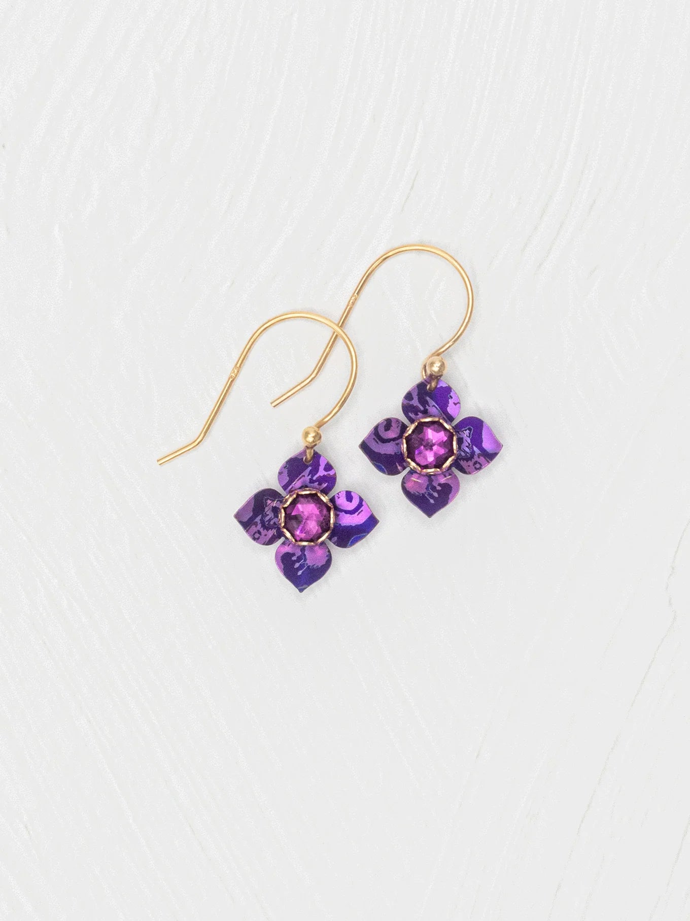 The Holly Yashi Petite Fleur Royal Purple Earrings feature gold hooks with purple flower-shaped pendants and faceted Austrian crystal centers, shown on a white textured background.
