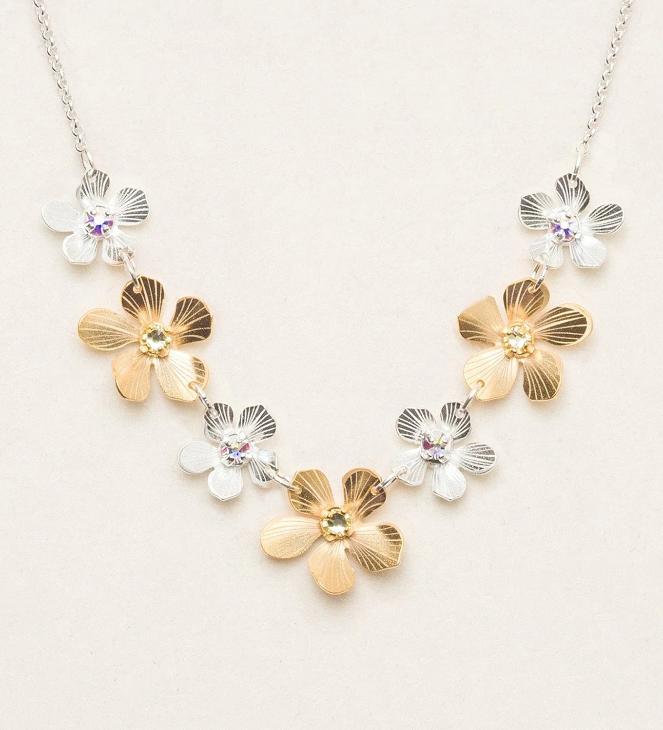 The Plumeria Classic Silver/Gold Necklace by Holly Yashi features alternating gold and silver flower-shaped charms with gemstone centers, beautifully displayed on a light beige background.