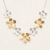 The Plumeria Classic Silver/Gold Necklace by Holly Yashi features alternating gold and silver flower-shaped charms with gemstone centers, beautifully displayed on a light beige background.