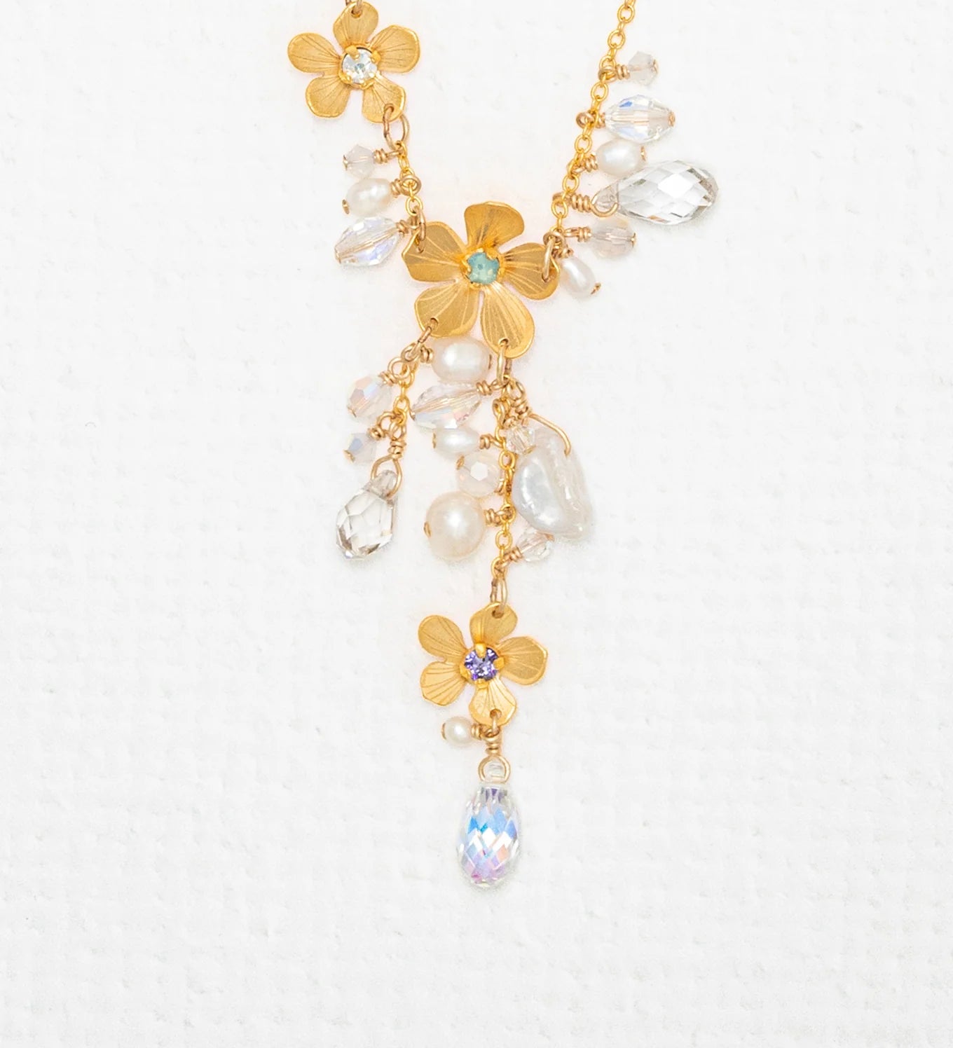 The Rafaela Gold & White Necklace by Holly Yashi features 18kt gold with plumeria flower pendants and sparkling clear and iridescent crystal beads, showcased on a textured white background.