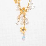 The Rafaela Gold & White Necklace by Holly Yashi features 18kt gold with plumeria flower pendants and sparkling clear and iridescent crystal beads, showcased on a textured white background.