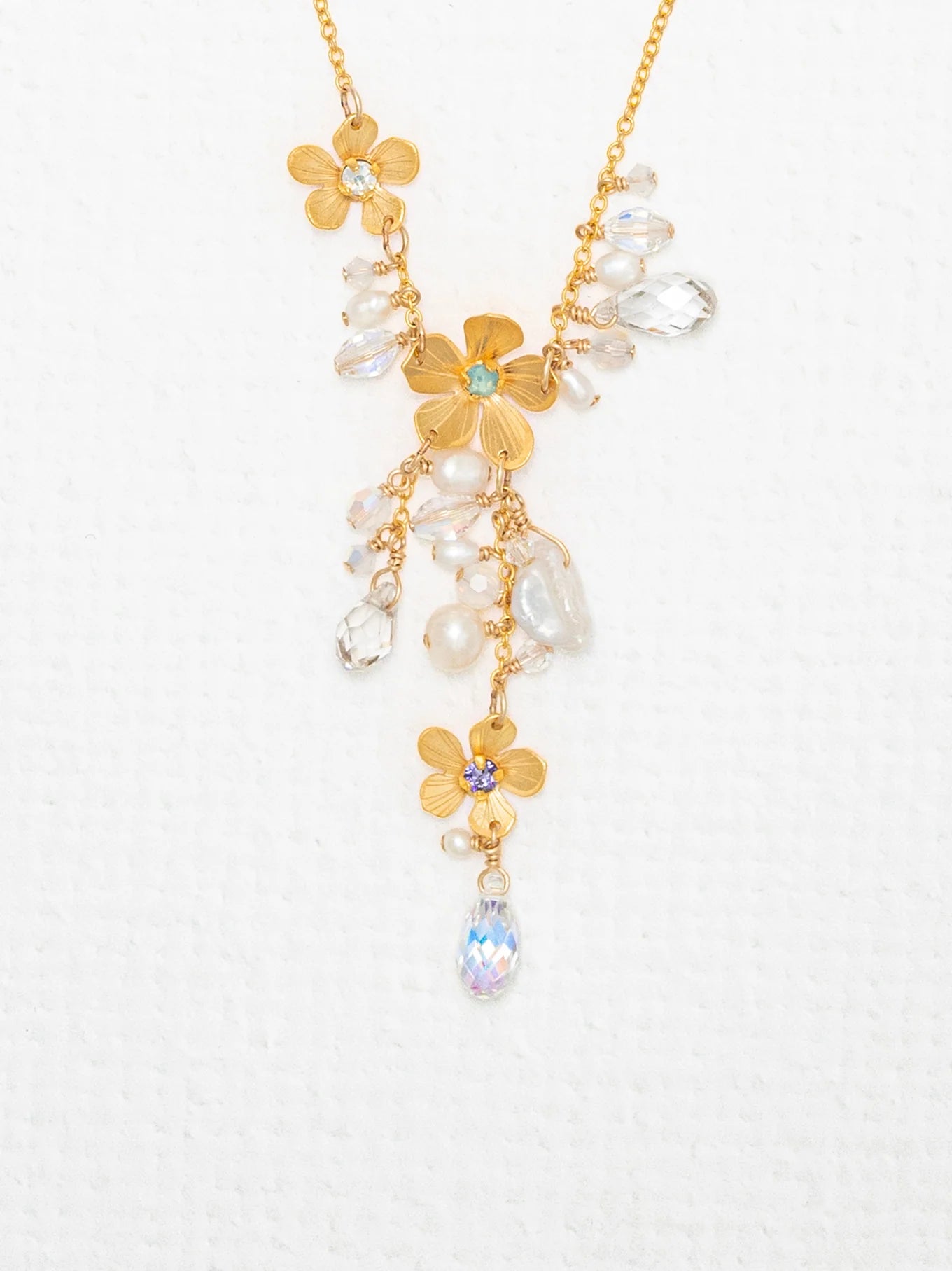The Rafaela Gold & White Necklace by Holly Yashi features 18kt gold with plumeria flower pendants and sparkling clear and iridescent crystal beads, showcased on a textured white background.