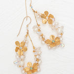 The Rafaela Gold & White Earrings by Holly Yashi feature gold-plated floral designs with delicate petals, small white pearls, and clear beads, shown on a white textured background.